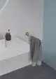 Handtuchstange in Brushed Gold mit grauem Handtuch hängt an einer wandmontierten Halterung in einem modernen Badezimmer von Ideal Standard. Minimalistische Einrichtungselemente sind sichtbar.
